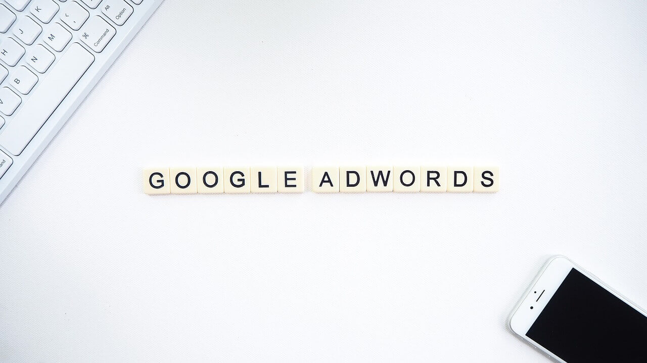 The words *Google AdWords* displayed on a white table. Part of a keyboard is visible on one corner of the table and a cell phone on the other.