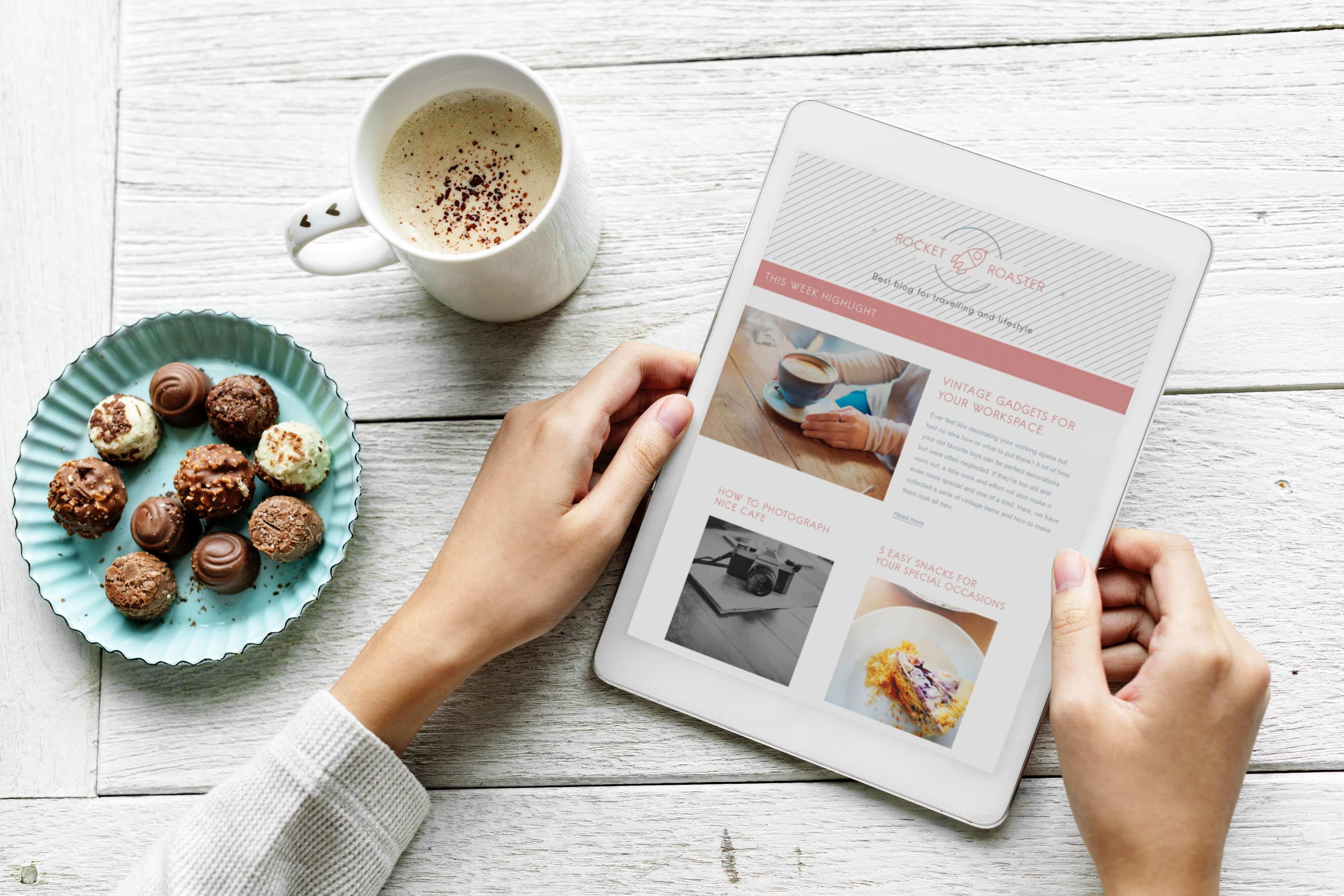 A white table with a cup of coffee and a plate of sweets placed over it. A pair of female hands are carrying a tablet showing some blog posts.