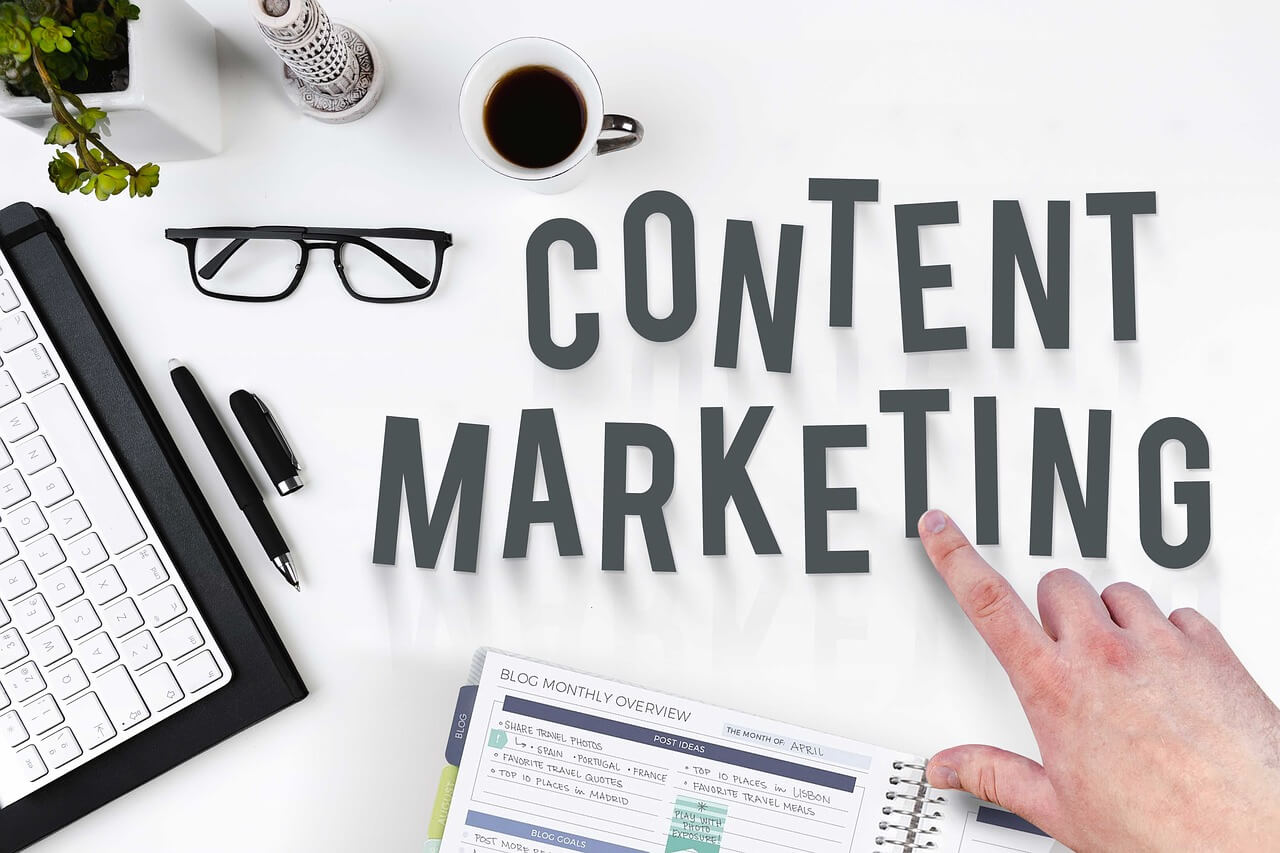 A white table top with some work essentials placed over it with the words *Content Marketing* that a finger is pointing at.